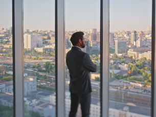 A man in a suit stands by a large window, gazing at the city skyline at sunset. The mood is contemplative and calm.