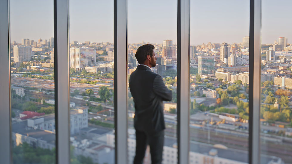 A man in a suit stands by a large window, gazing at the city skyline at sunset. The mood is contemplative and calm.