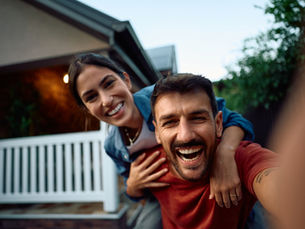 A couple laughs joyfully in the front yard, with one giving the other a piggyback ride. A white picket fence and house are in the background.