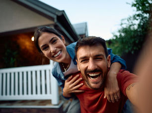 A couple laughs joyfully in the front yard, with one giving the other a piggyback ride. A white picket fence and house are in the background.