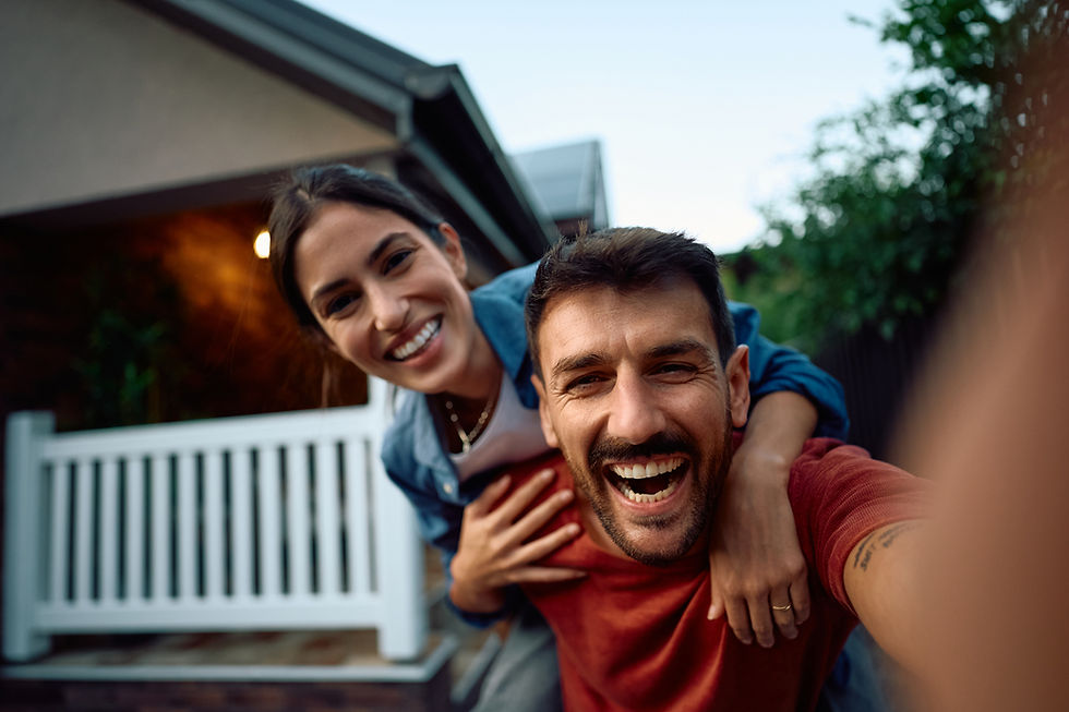 A couple laughs joyfully in the front yard, with one giving the other a piggyback ride. A white picket fence and house are in the background.