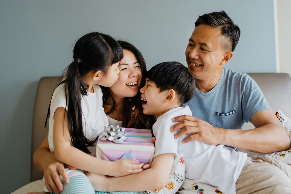 Family of four laughing together on a sofa. A girl kisses her mom while the son holds a gift box with a silver bow. A joyful, cozy scene.