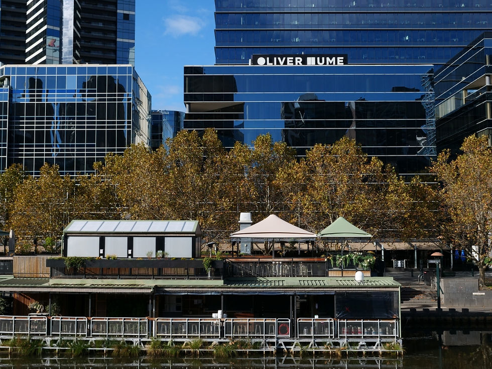 Blue skyscraper with reflections, text "OLIVER HUME" on top. Autumn trees and riverside buildings in foreground. Clear sky above.