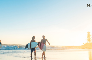 Four surfers walking out of the water with wet hair, holding surf boards as the sun sets in the background. - with text overlayed saying "no surpise that the Reserve Bank of Australia held interest rates.