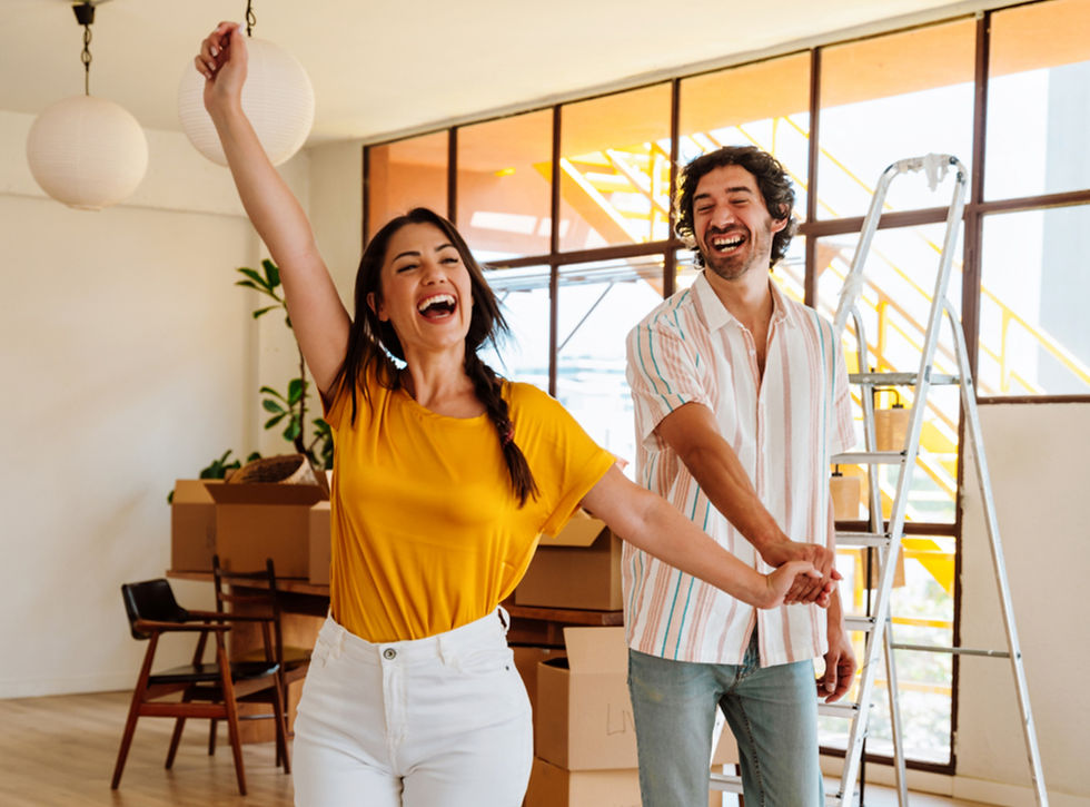 A joyful couple stands in a bright room, surrounded by moving boxes and a ladder. The woman in yellow raises her arm, smiling widely.